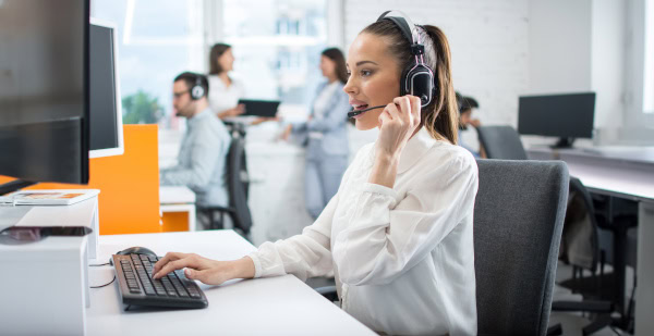 Eine Frau im Büro mit Headset tippt konzentriert auf einem Computer, im Hintergrund Kollegen.